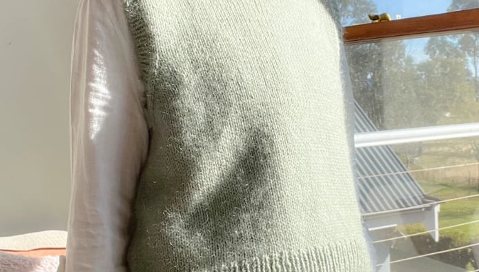 Woman wearing a dusty green hand knit slipover with a white shirt and jeans in a sunny room in front of a window. 