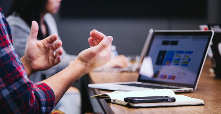 A businessman gesturing during a collaborative office meeting with a laptop and notebook on the table.