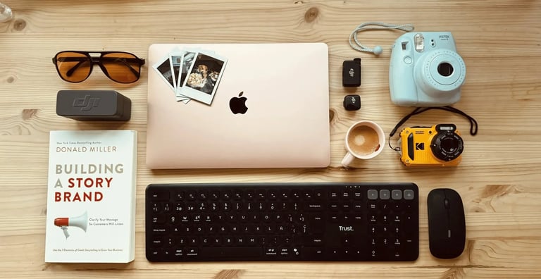 Flat lay of a workspace with a MacBook laptop, Instax camera, Trust keyboard, and Building a Storybrand book.