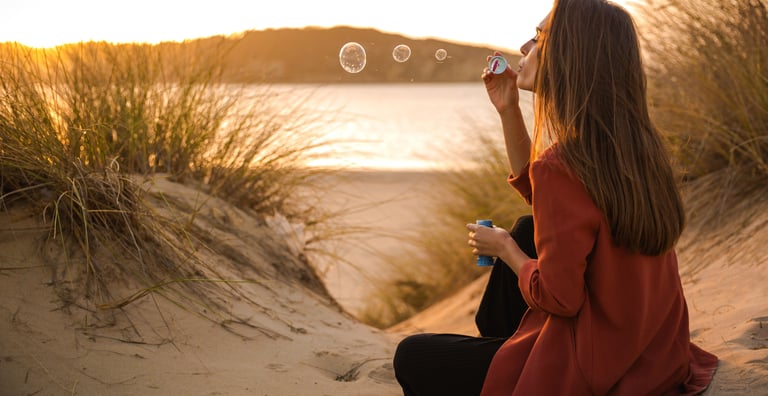 Mujer de mediana edad sentada en la arena en el atardecer mientas hace burbujas de jabón tranquilamente, paz mental