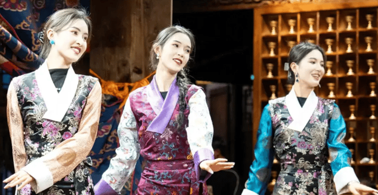 Three women in traditional Tibetan silk dresses perform a cultural dance on a stage.