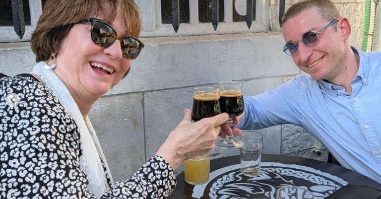A smiling man and woman toast with dark craft beers at an outdoor pub table.