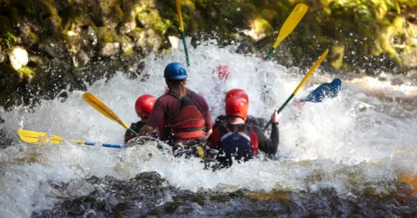 A group of people wearing helmets white water rafting through splashing river rapids with yellow paddles.