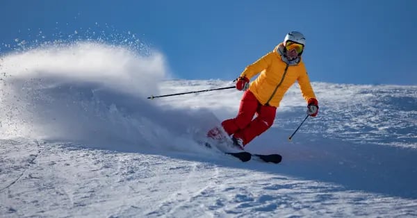 A skier in a yellow jacket and red pants carves through fresh powder on a sunny mountain slope.
