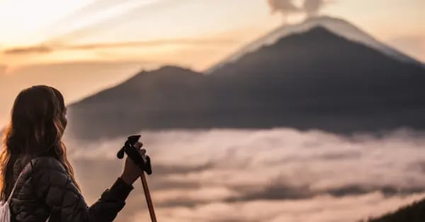 A hiker with trekking poles overlooks Mount Batur volcano at sunrise above the clouds in Bali.