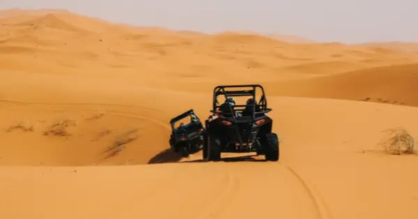 Adventure seekers driving black off-road buggy vehicles across golden Sahara Desert sand dunes.