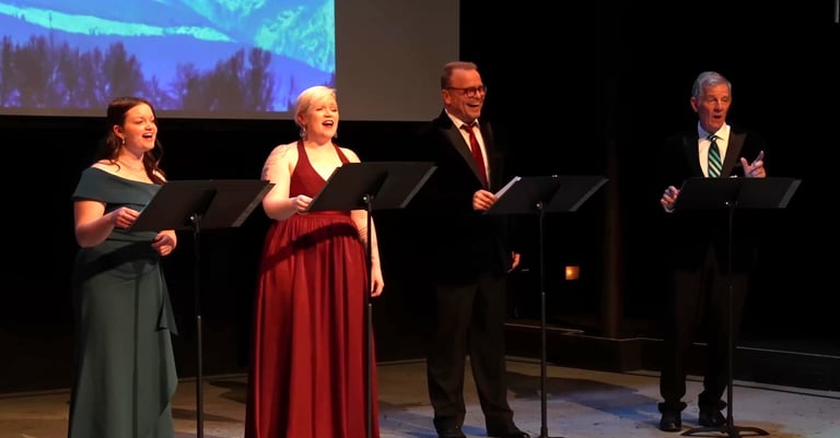 Four singers in formal attire perform a vocal concert on stage with music stands.