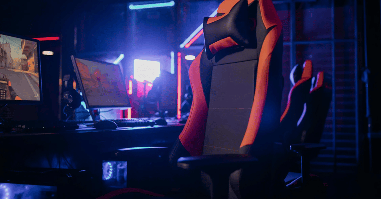 silla gaming color negro con naranja y en el fondo una pc gamer con monitor y diadema
