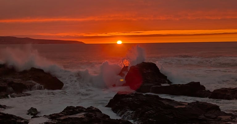 Golden sunset over the ocean with waves crashing against rocky coastline under an orange sky.