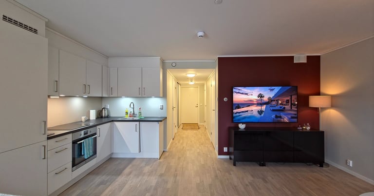 Modern open plan apartment featuring a white kitchen and living area with a wall-mounted TV on a red accent wall.