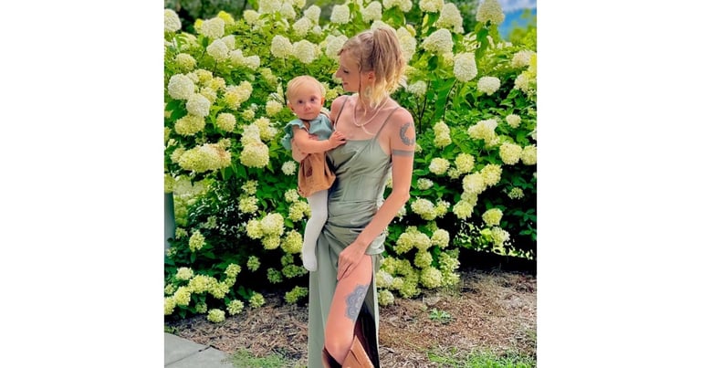Jade in a green dress and boots holding baby Nova by flow bush, representing maternal instinct and caregiving
