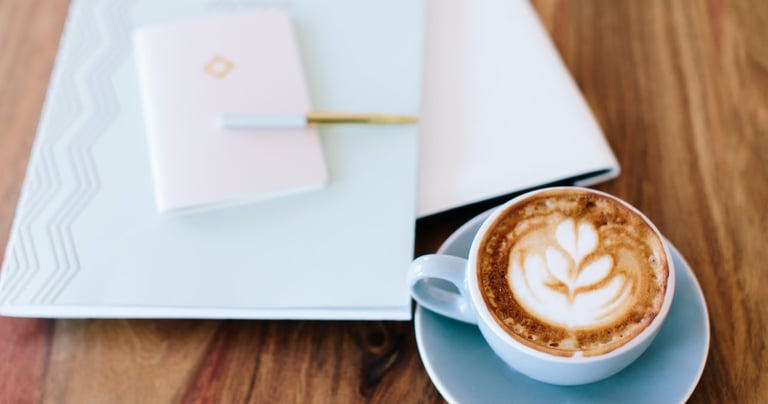 A cup of latte with foam art sits on a saucer next to a closed notebook with a pen on a wooden table