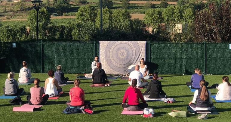 An outdoor yoga class practicing on a green lawn with a scenic mountain backdrop and mandala tapestry.