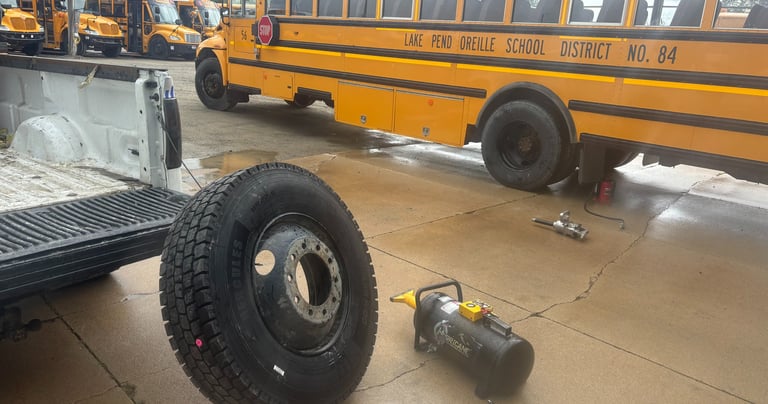 A mechanic services a yellow school bus tire and wheel on a wet pavement lot.