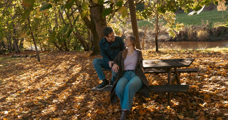 An engage couple sitting on a wooden picnic table in a park during autumn with golden fallen leaves.