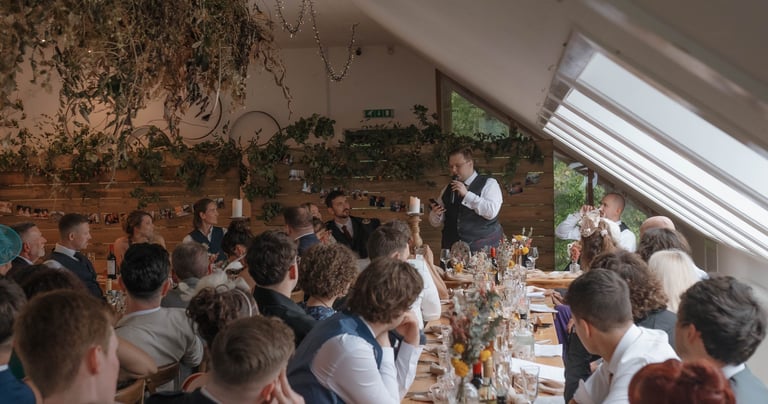A best man in a kilt gives a wedding toast to guests at a rustic indoor reception venue.