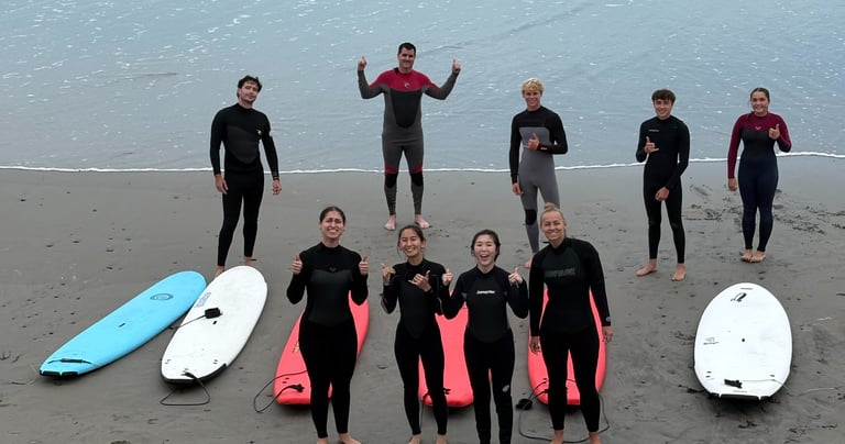 A group of surfers in wetsuits posing with surfboards on a sandy beach after a surfing lesson.