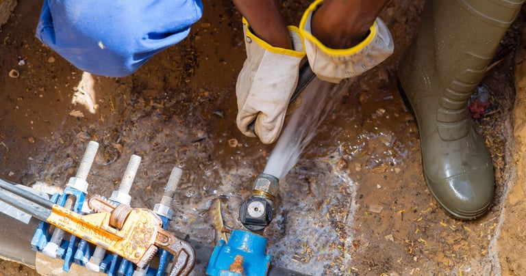 Plombier réparant à la clé un tuyau d'eau enterré dans une tranchée boueuse.