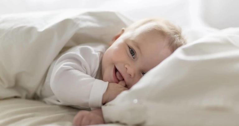 A happy, smiling newborn baby boy lying in bed under a white duvet cover.