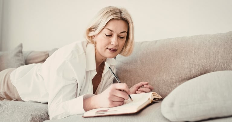 Blonde woman lying on a grey sofa writing in her journal or planner with a pen.