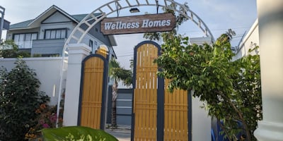 A decorative archway with a wooden sign reading 'Wellness Homes' stands above yellow gates. Lush gre