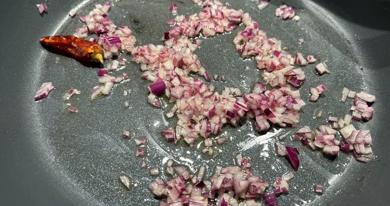 Finely diced red onions and a whole dried chili pepper sautéing in oil in a non-stick frying pan.