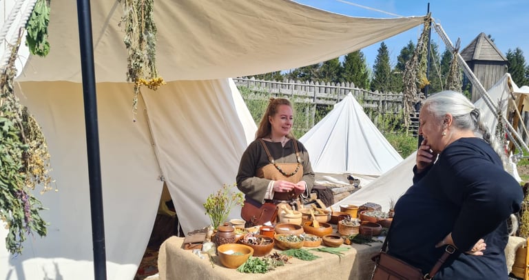 a woman dressed in viking clothing standing behind a table of herbal remedies talking  with visitors