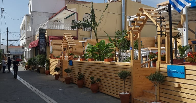 Greek tavern outdoor seating area with wooden fencing, potted plants, and a Greek flag on a cobblestone street.