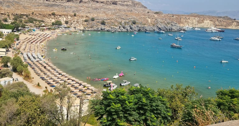 Panoramic view of Lindos beach in Rhodes with turquoise water, rows of umbrellas, and boats at anchor.
