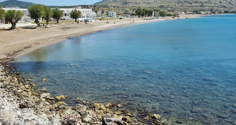 Panoramic view of a sandy Greek beach with turquoise sea, white buildings, and coastal mountains under a clear blue sky.