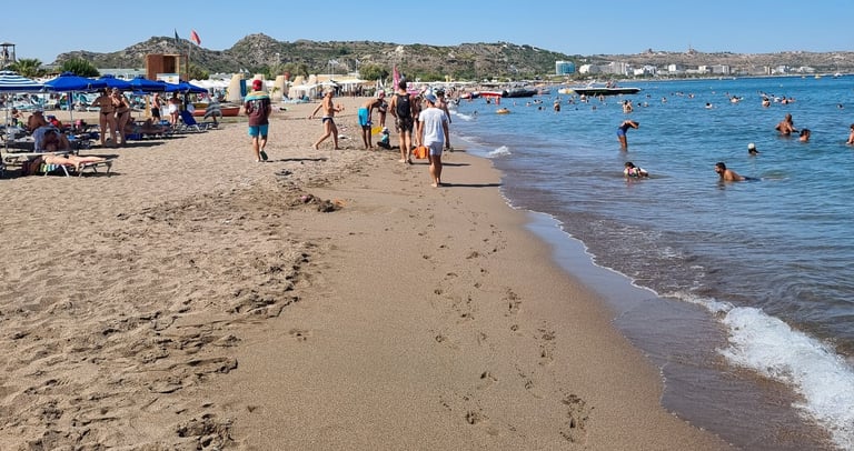 Crowded sandy beach in Rhodes, Greece, with tourists swimming in the blue Mediterranean Sea under a clear sky.