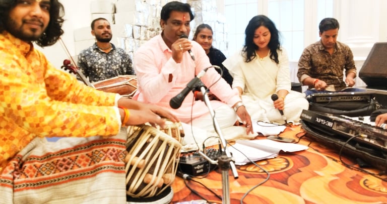 Indian musicians performing a devotional bhajan with tabla drums, harmonium, and vocal microphones.
