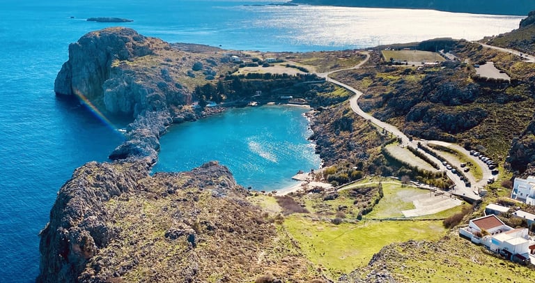 Aerial view of St. Paul's Bay in Lindos, Rhodes, featuring turquoise water, rocky cliffs, and the Mediterranean Sea.