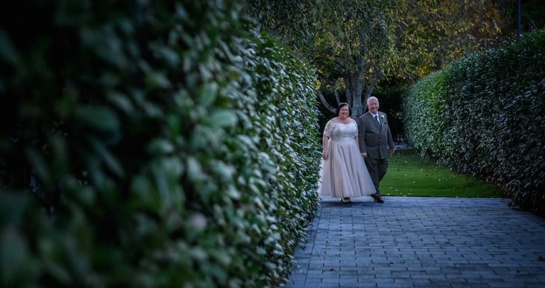 Smiling bride in white lace dress and groom in suit walking on stone path between green hedges.
