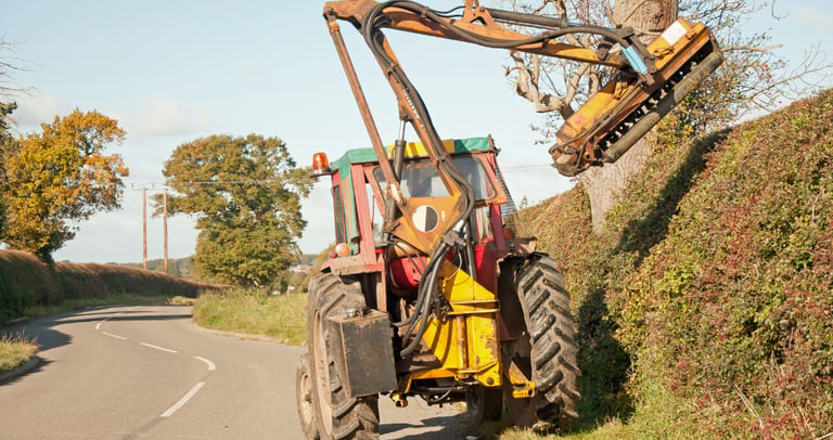 A tractor with a mechanical flail arm attachment trims a tall roadside hedge and trees.