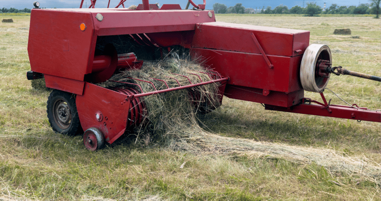 Red square hay baler harvesting dry grass in a rural field under a blue sky.