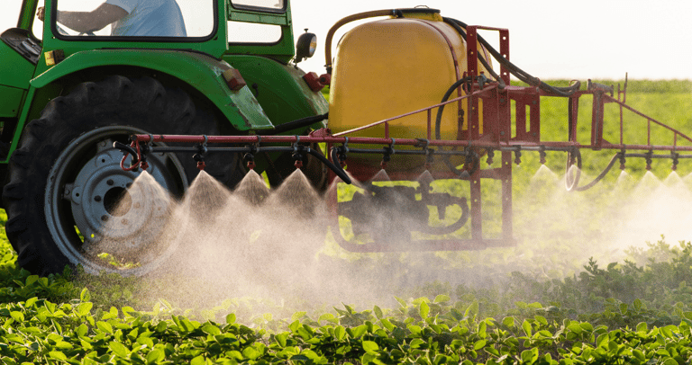 A green tractor with a yellow tank boom sprayer applying liquid fertilizer to a field of crops.