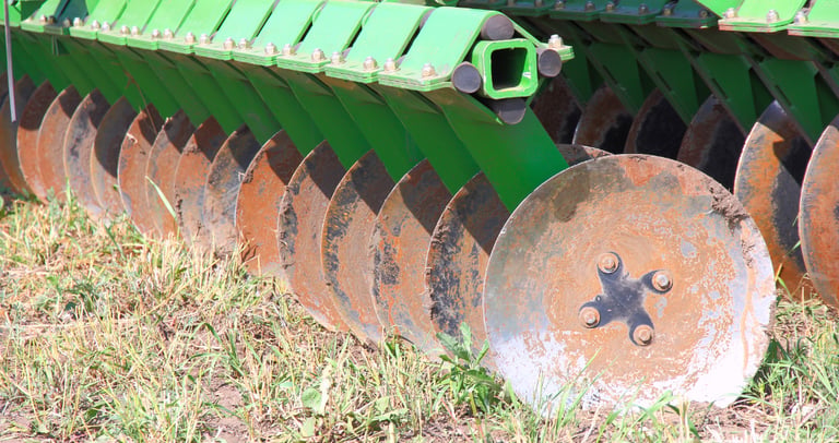 A green agricultural disc harrow with steel blades tilling dry soil in a farm field.