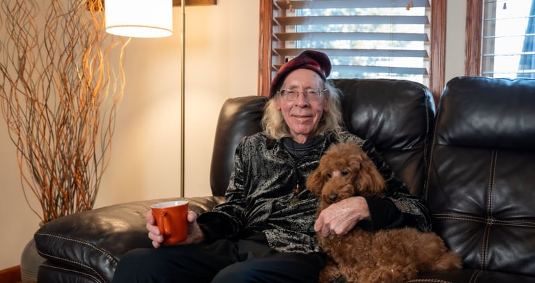 A senior man wearing a beret sitting on a leather sofa with a brown toy poodle and a coffee mug.