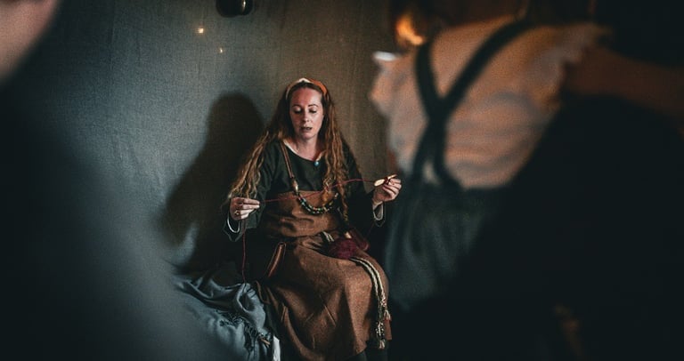 A woman in a Viking age dress sitting on a bench using a spindle