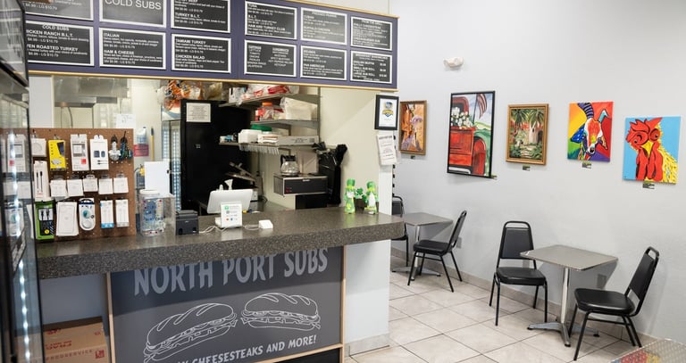 Interior of North Port Subs sandwich shop featuring a service counter, menu board, and local art.