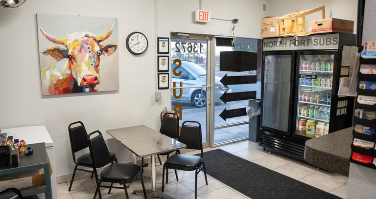 Indoor seating area at North Port Subs featuring a colorful cow painting, dining table, and beverage cooler.
