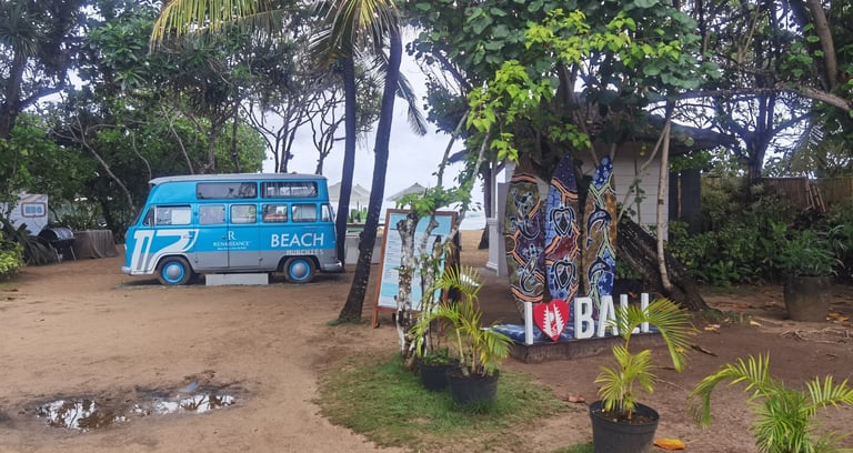 A vintage blue food truck parked near tropical palm trees and surfboards at a Bali beach resort.