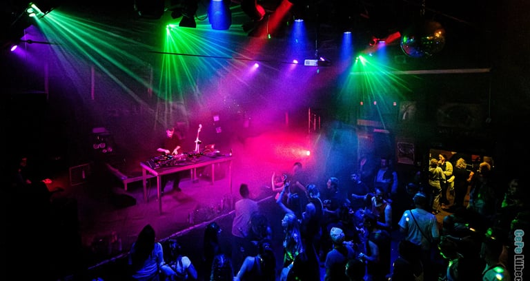 A DJ performs on a dark stage with vibrant neon laser lights and a dancing crowd at a nightclub event.