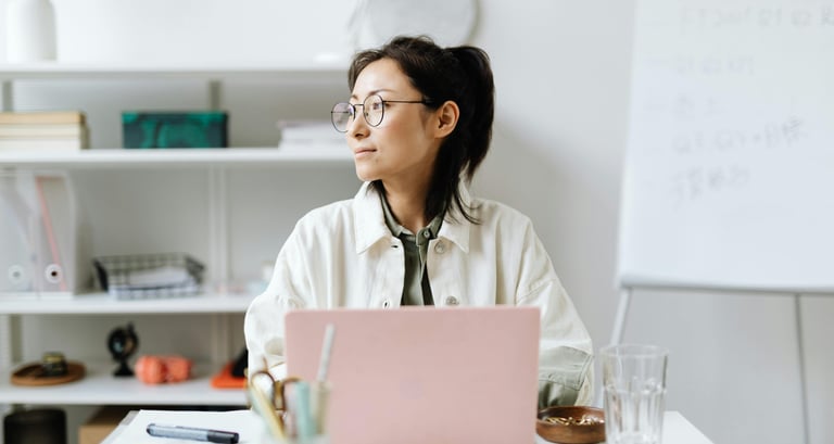 Professional woman with glasses working on a pink laptop in a modern, bright office setting.