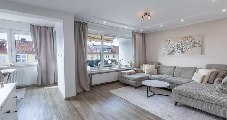 Modernes Wohnzimmer mit grauem Ecksofa, Holzboden und großen Fenstern mit neutralen Vorhängen.