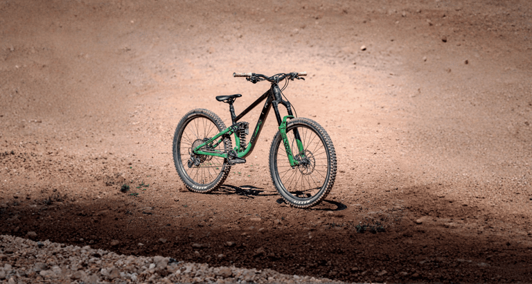 A green and black full-suspension mountain bike parked on a dirt trail at a bike park.