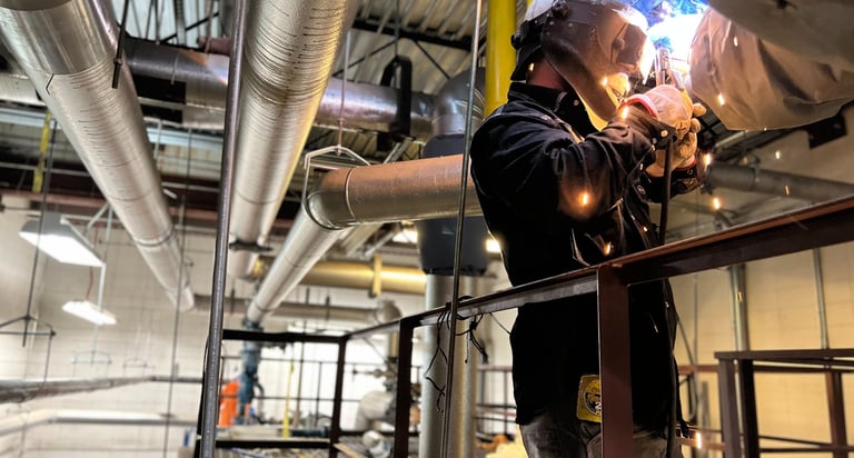 Industrial welder in protective gear performing precision pipe welding in a factory setting.