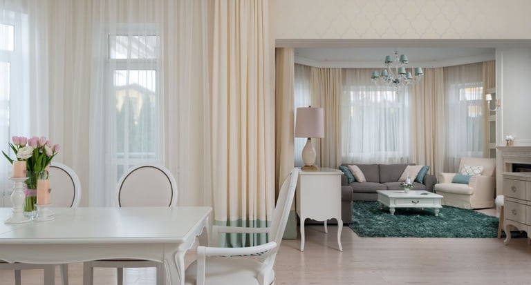 Elegant open concept living and dining room featuring white furniture, soft cream curtains, and a teal area rug.