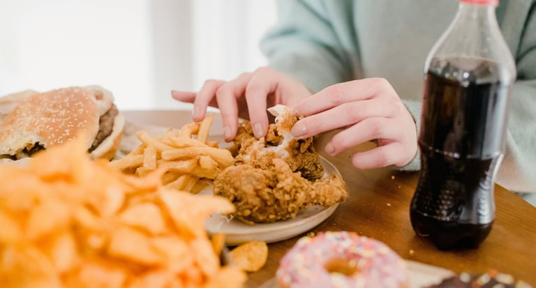 Person eating fried chicken with a side of french fries, burgers, donuts, and a cola soda.